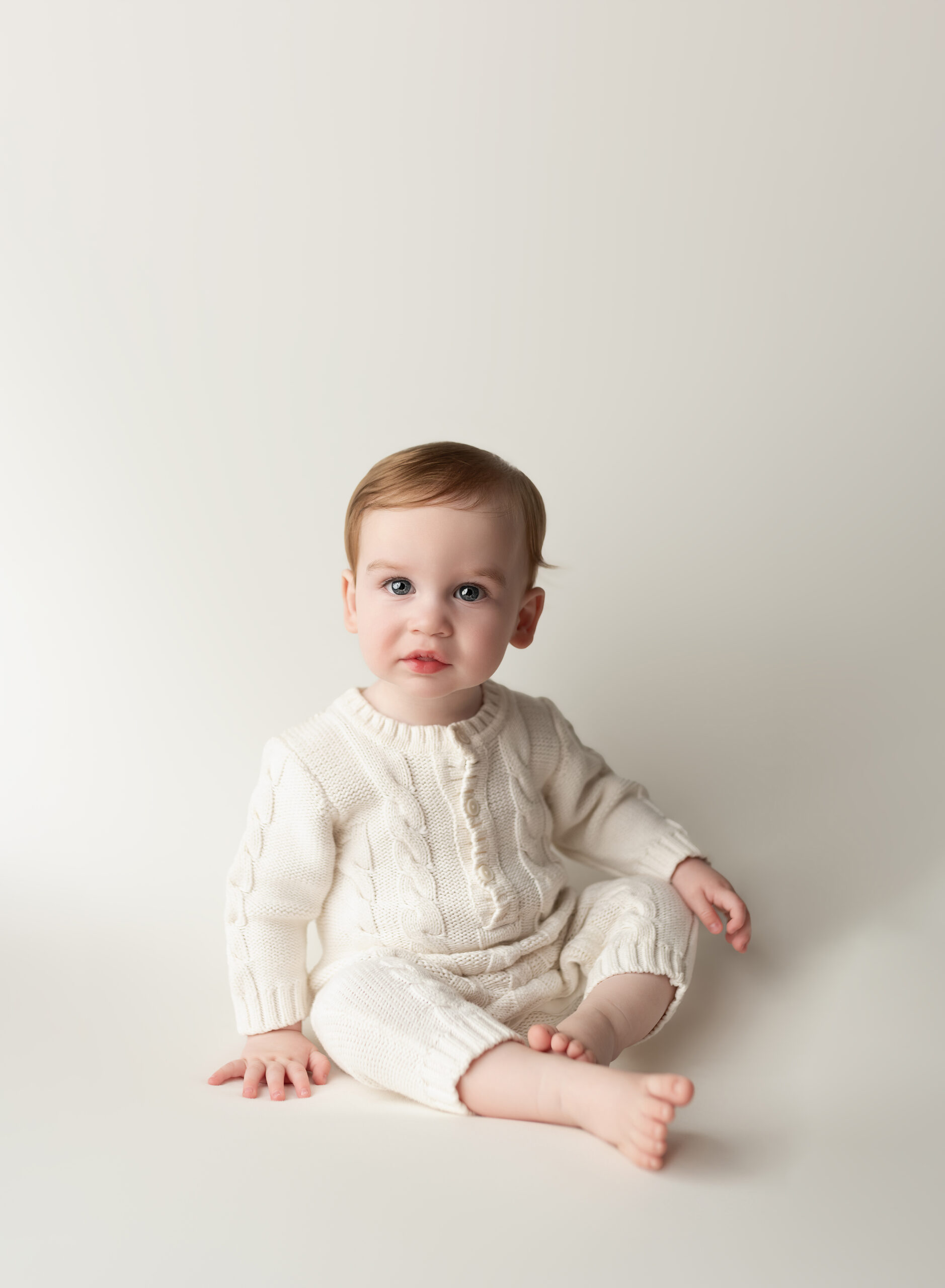 Baby sitting in a photography studio in Paris, Ontario, wearing a cream-colored knitted outfit, photographed by Meraki Photographic Creations, specializing in newborn, milestone, and family photography.