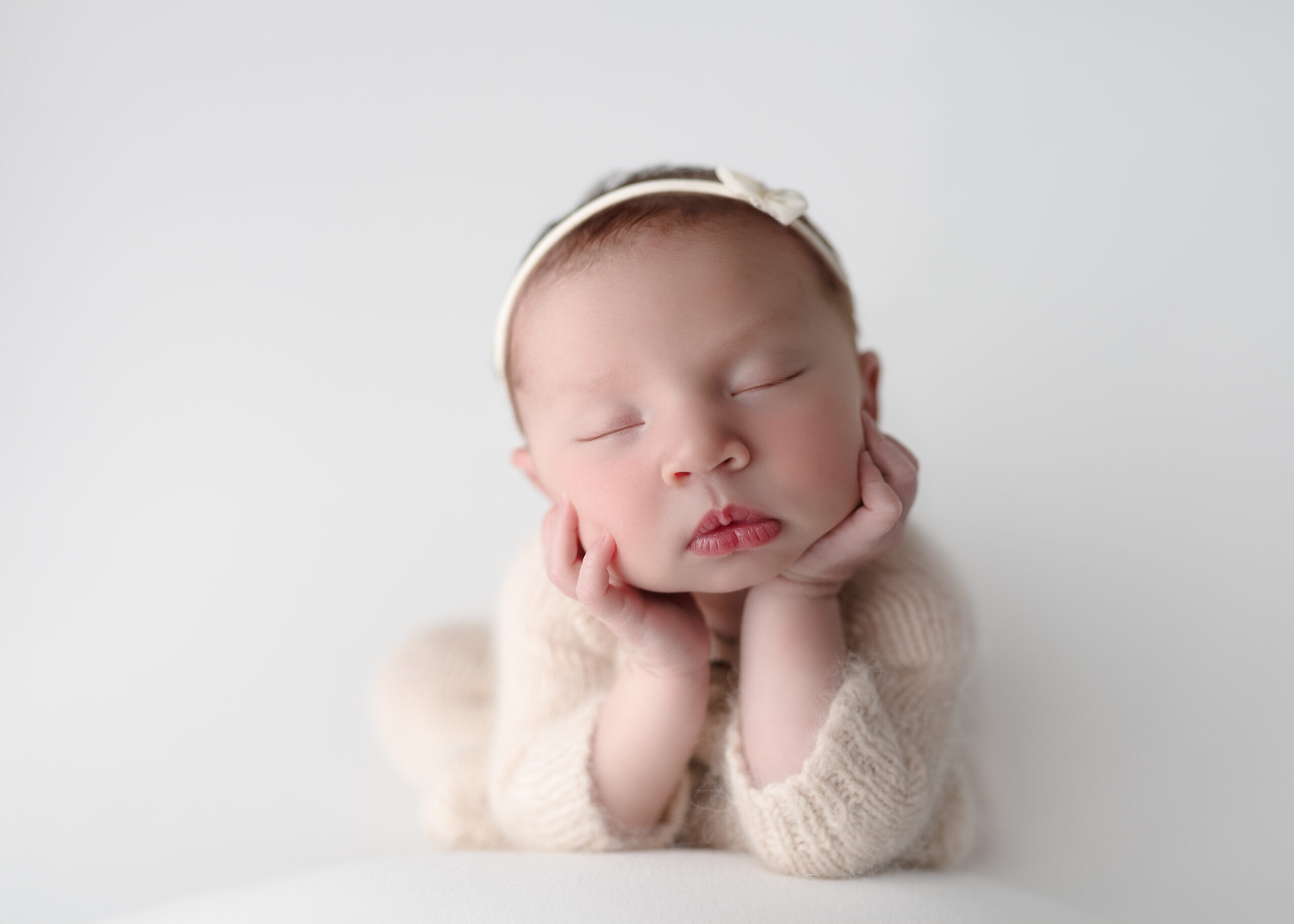 Newborn baby peacefully posed in a froggy position on a soft, neutral background, wearing a cream knitted outfit and a delicate headband, photographed by Meraki Photographic Creations in Paris, Ontario.