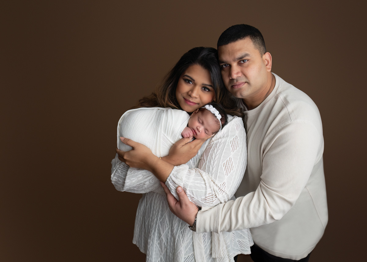 Family portrait with newborn baby in a warm-toned studio photography session in Paris, Ontario. Parents lovingly embrace their sleeping baby wrapped in a soft white swaddle.